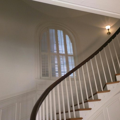White plantation shutters decorating arched window located in curved stairwell.